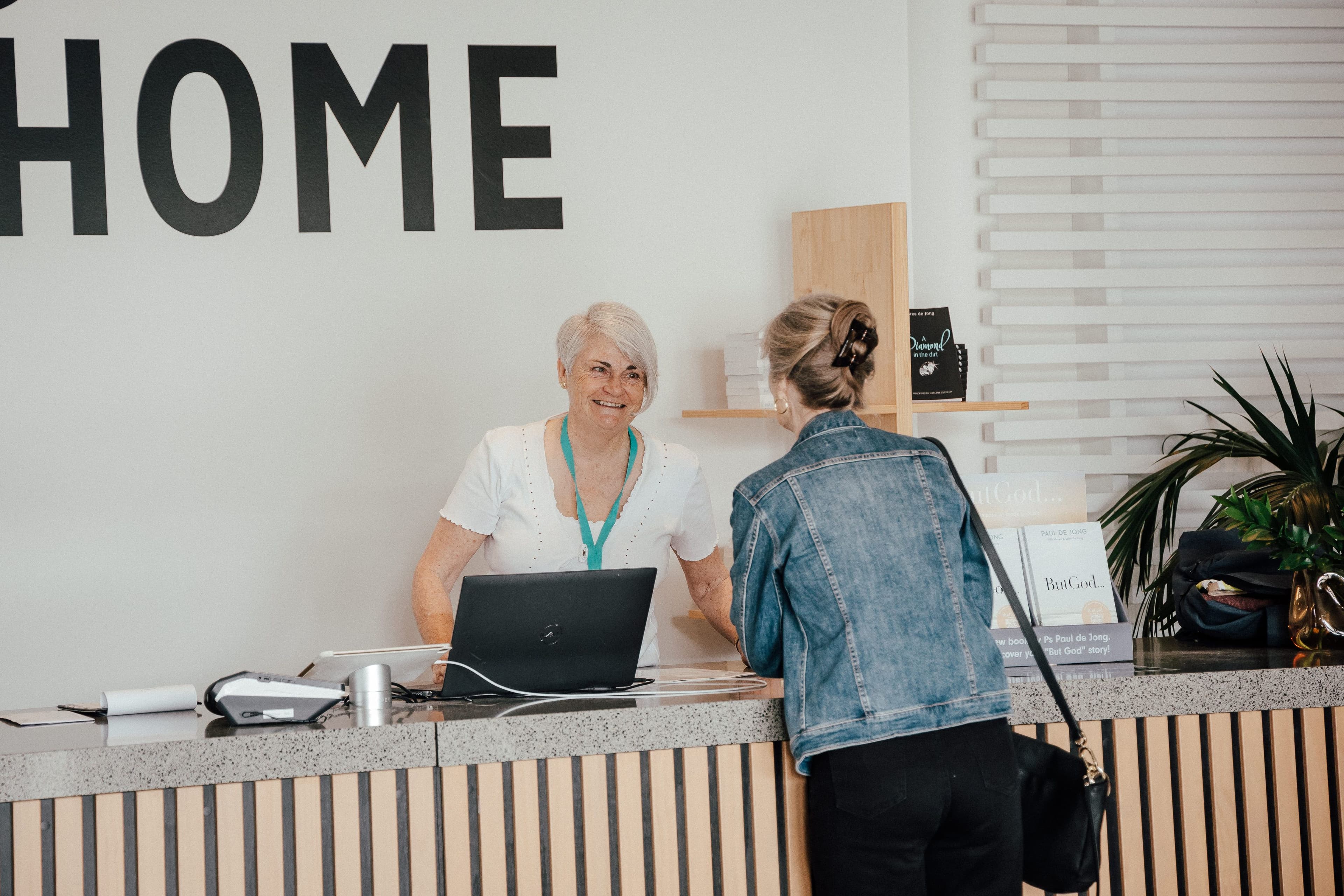 Woman behind the Info Desk and woman on the other side of the Info Desk talking to each other with WELCOME HOME on the wall behind them