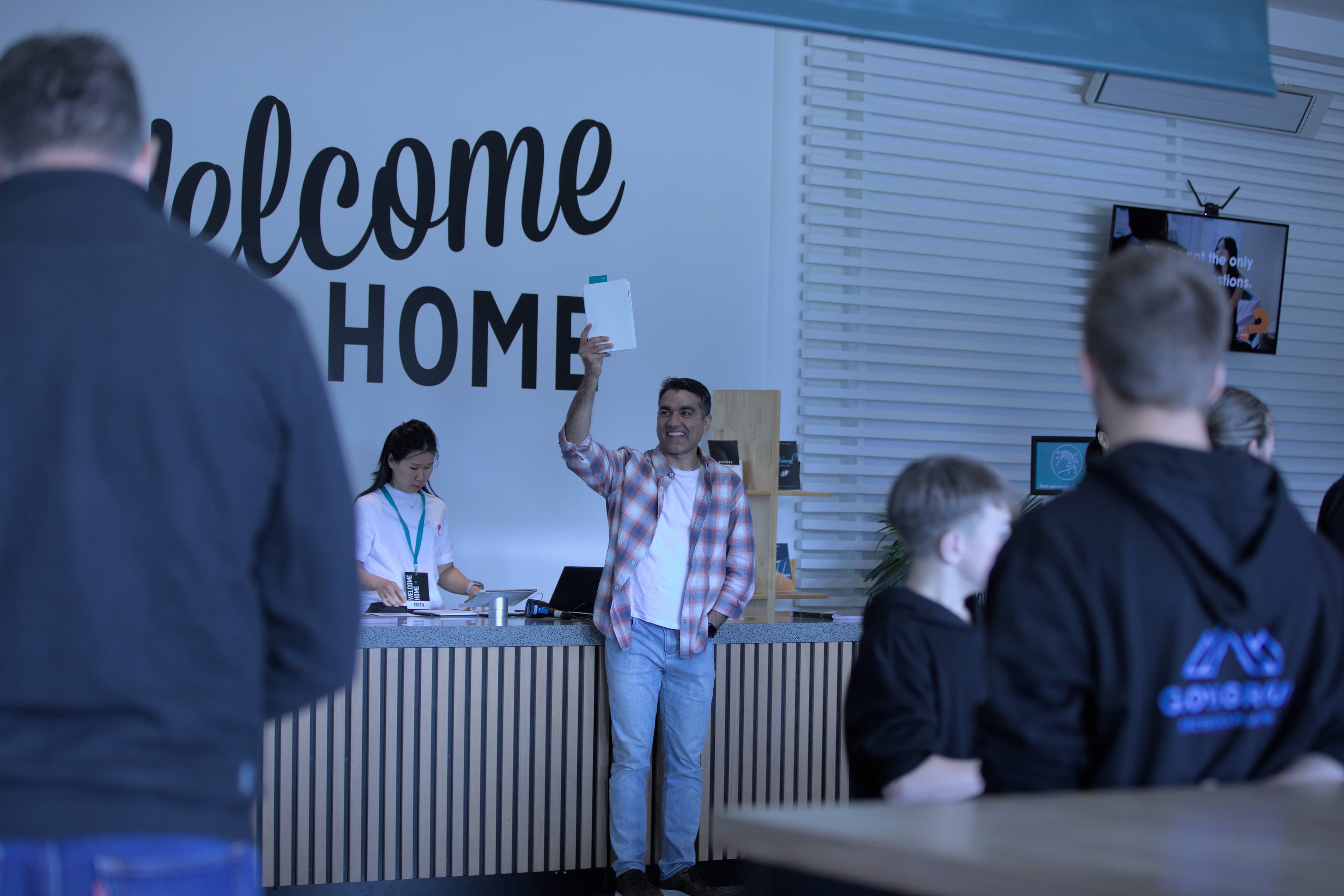 Man holding up a bible for new Christians in front of the Info Desk at North Campus with people around in the foyer and Welcome Home on the back wall