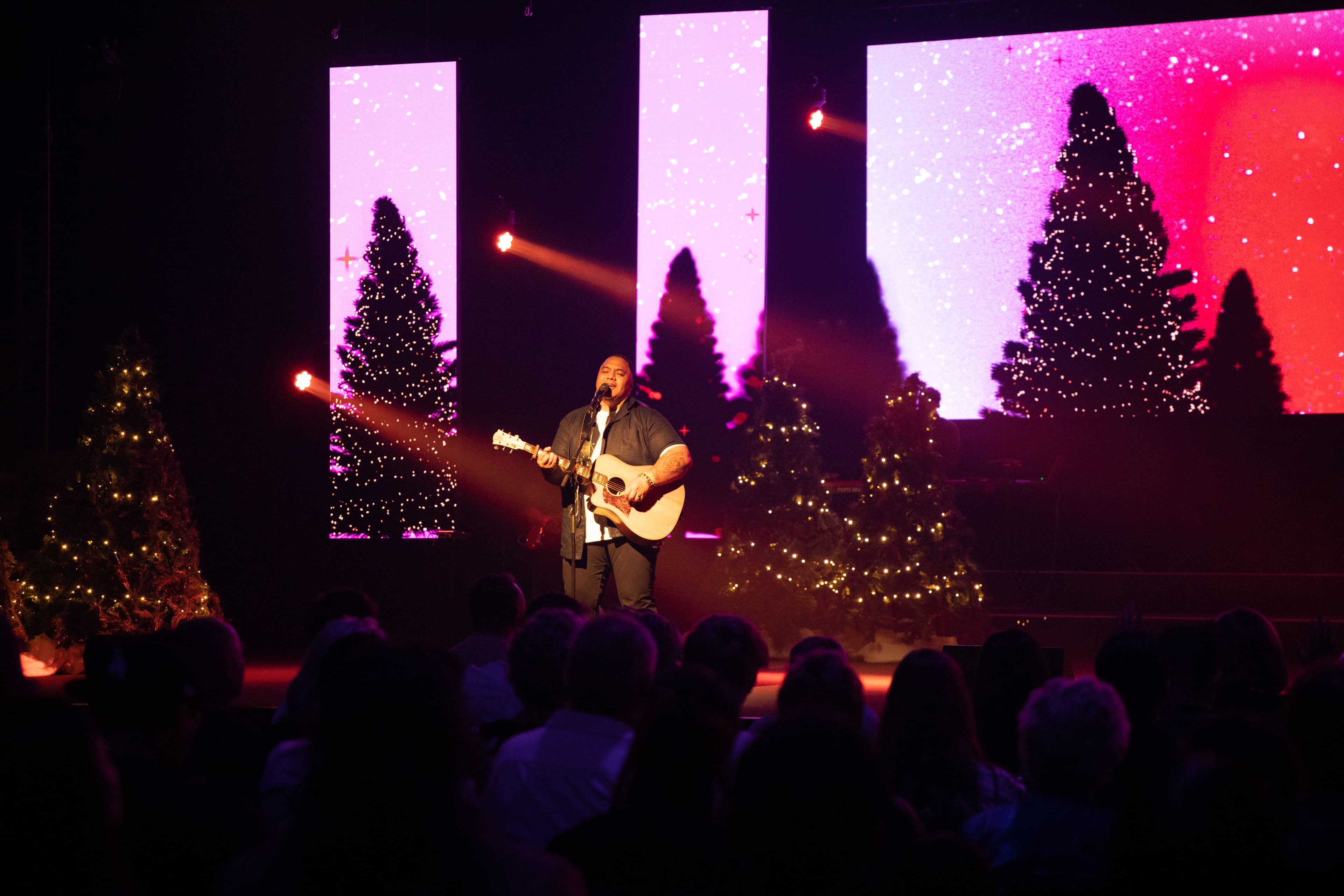Man standing on stage with a guitar singing with Christmas trees behind him