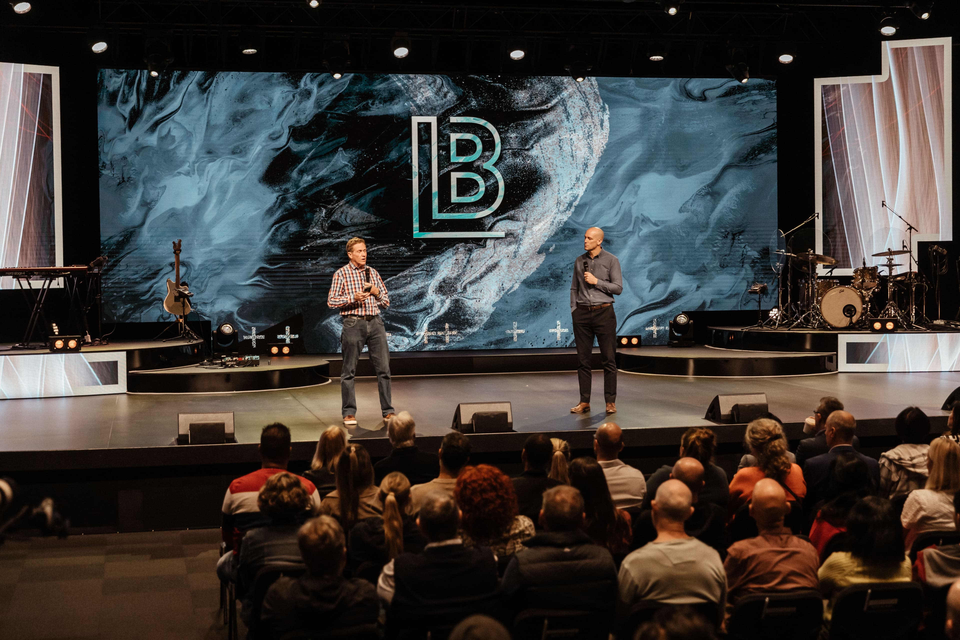 Two men standing on stage at Central Campus with the LIFE Business LB logo on screen behind them, discussing business at a LIFE Business event