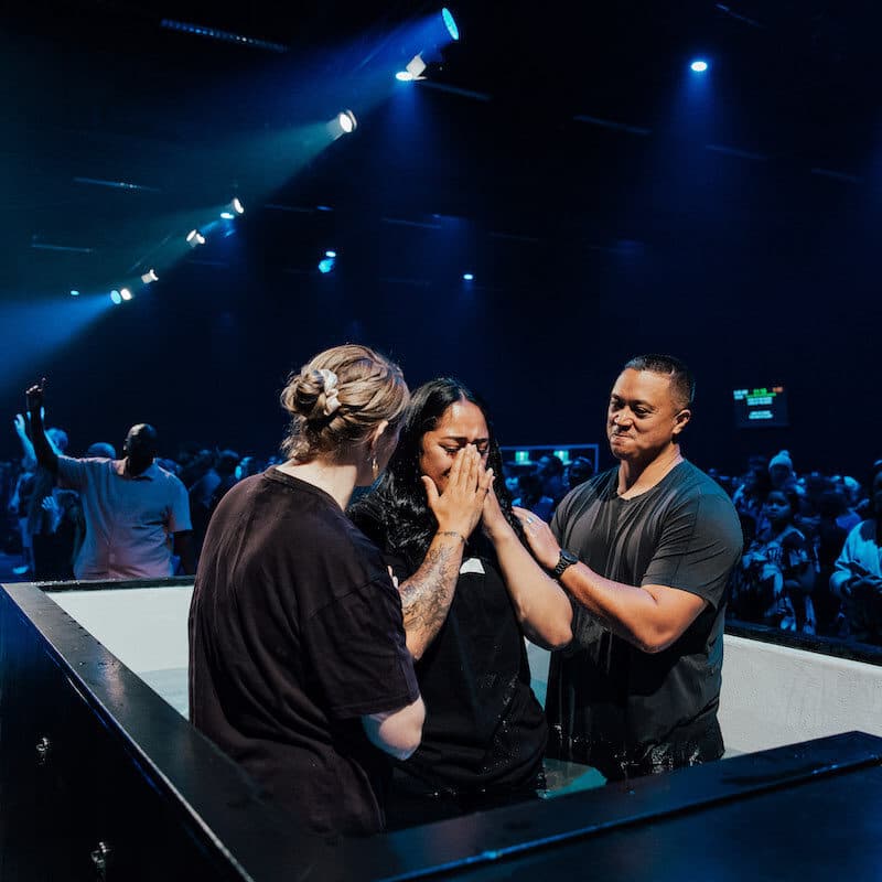 Person with hands over their face with emotion after being baptsed by two people while standing in baptism pool in LIFE South auditorium
