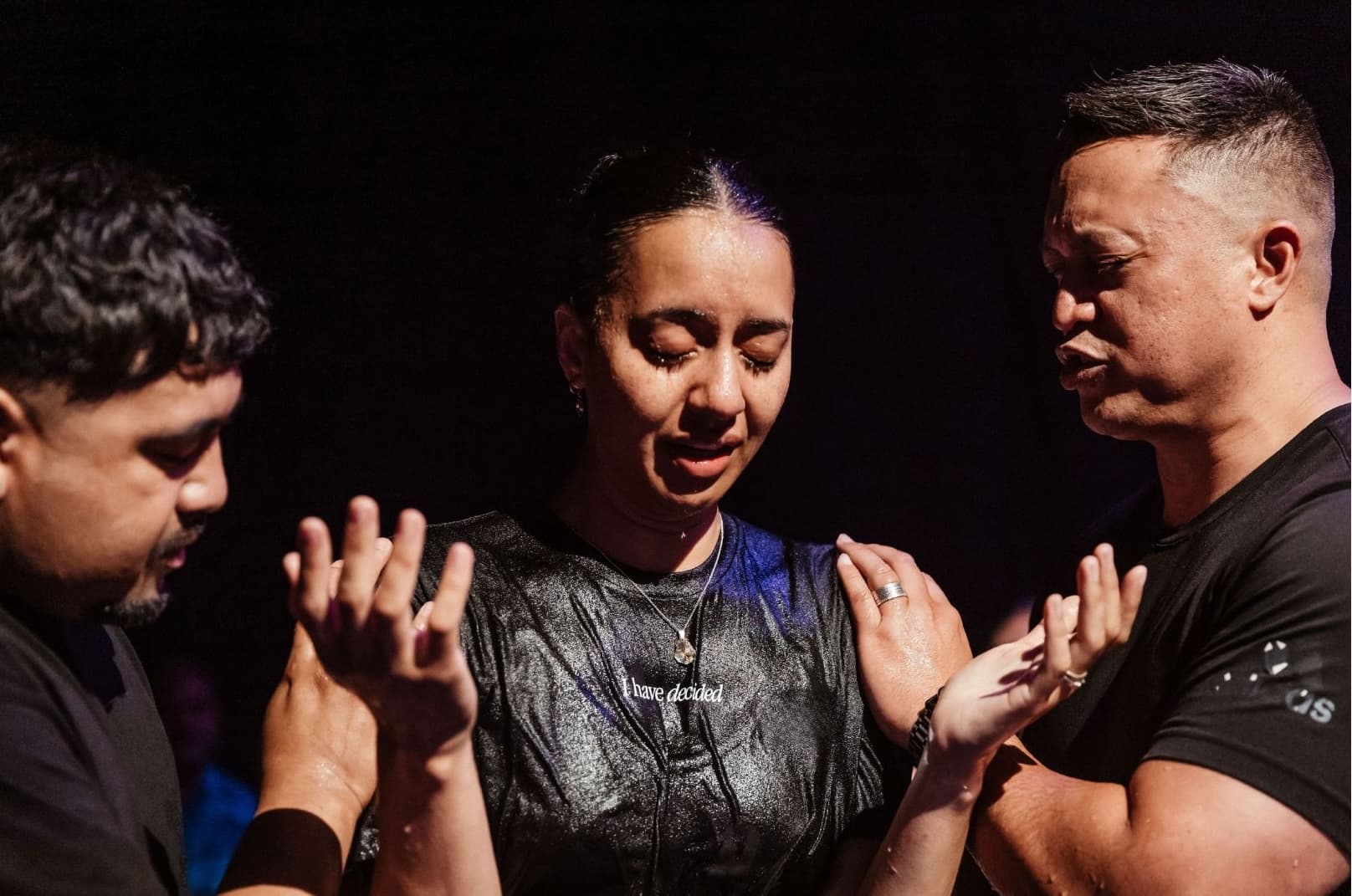 Woman who has just been baptised with hands raised in prayer being prayed for by a man either side of her in the baptism tank