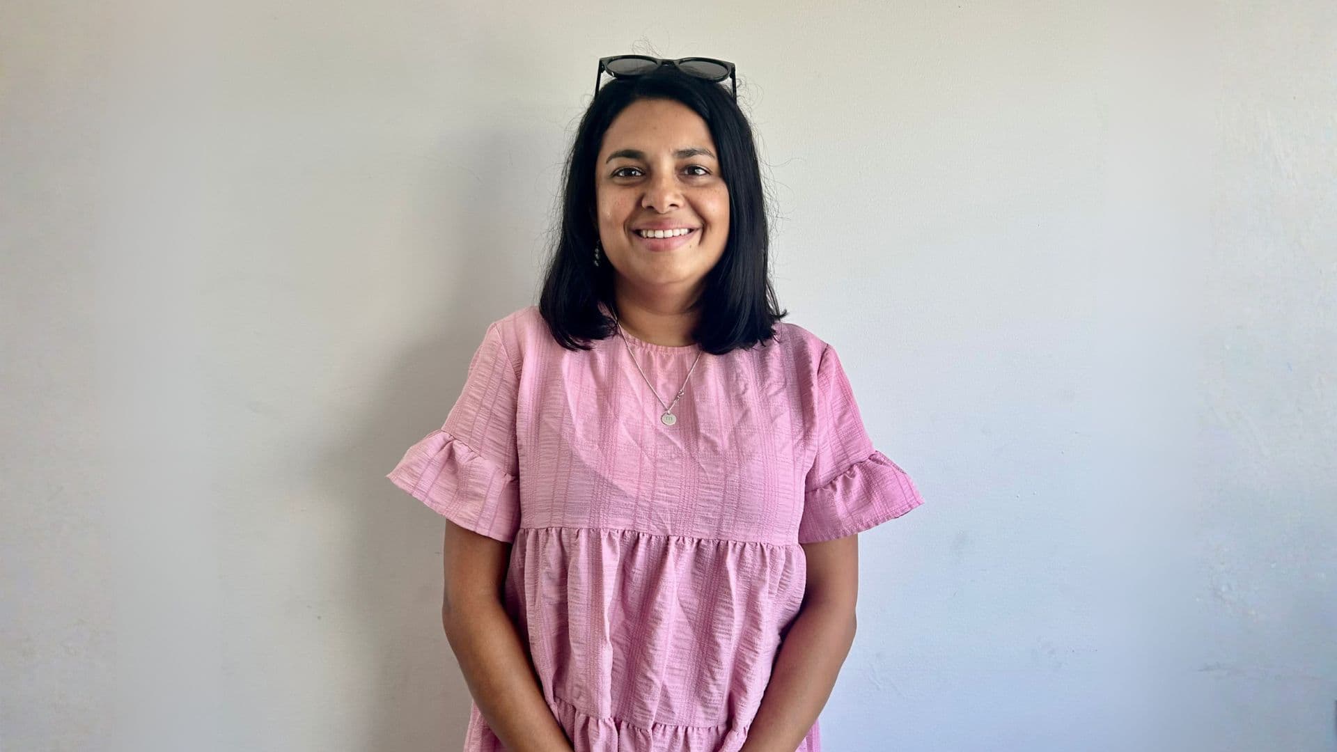 Lonelle standing against a pale grey background in a pink dress with sunglasses on her head
