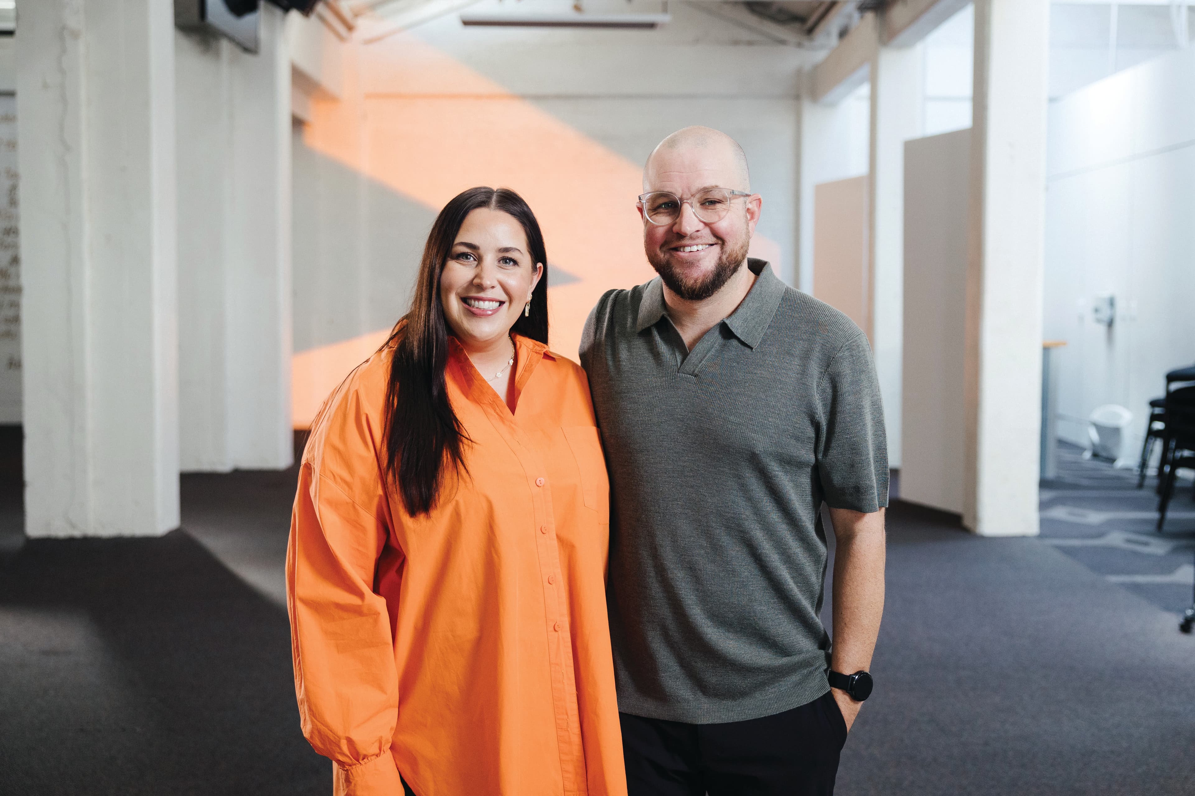 Ps Luke and Missy smiling at the camera with an orange > projected on the white wall behind them