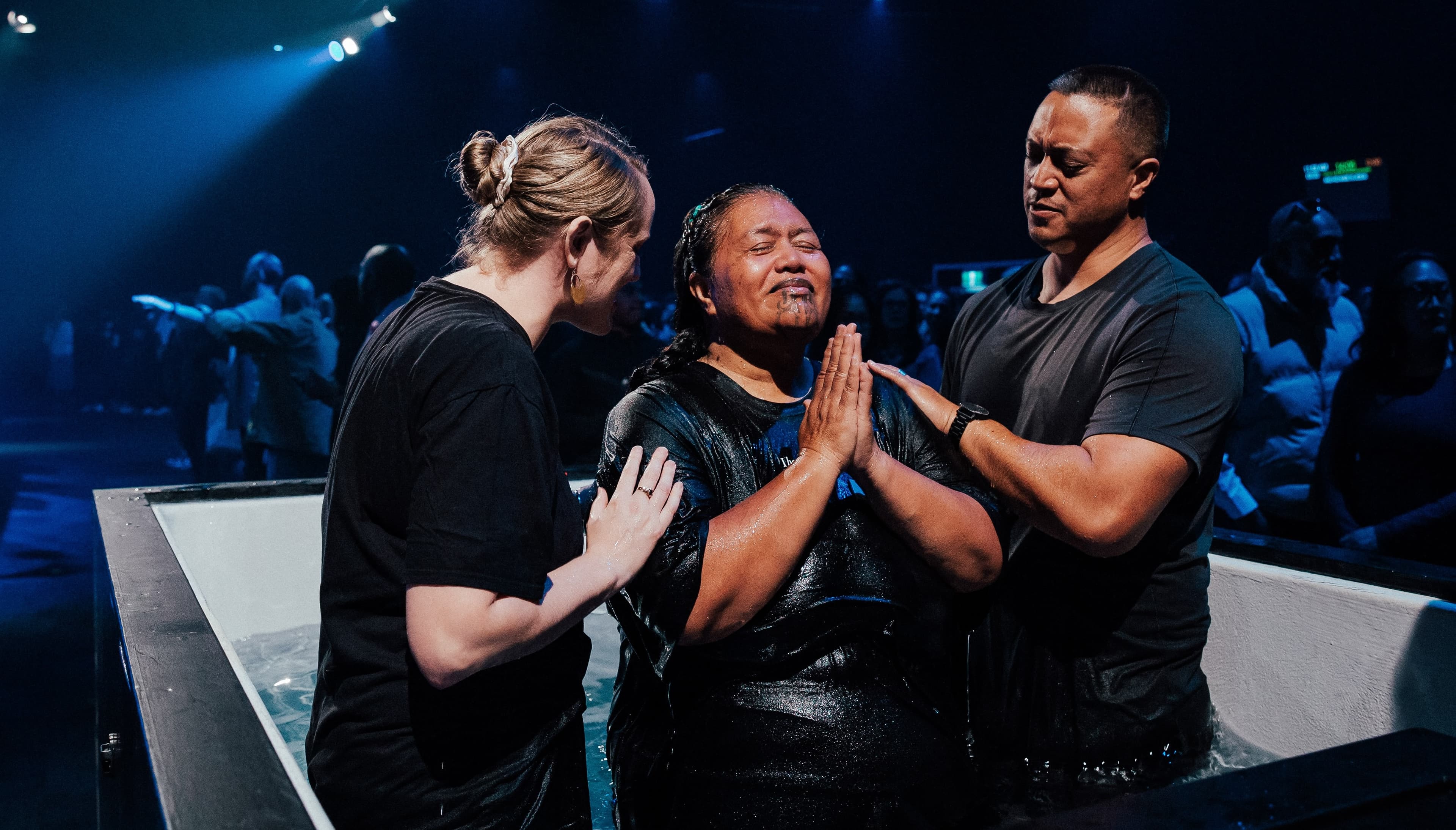 Woman who has just been baptised with her hands clasped together and eyes closed as she is prayed for by a woman on her left and man on her right