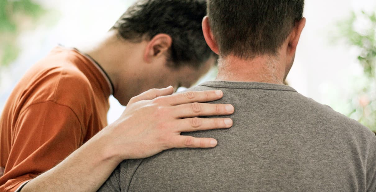 Two men from behind with one man laying a hand on another man's shoulder while they pray